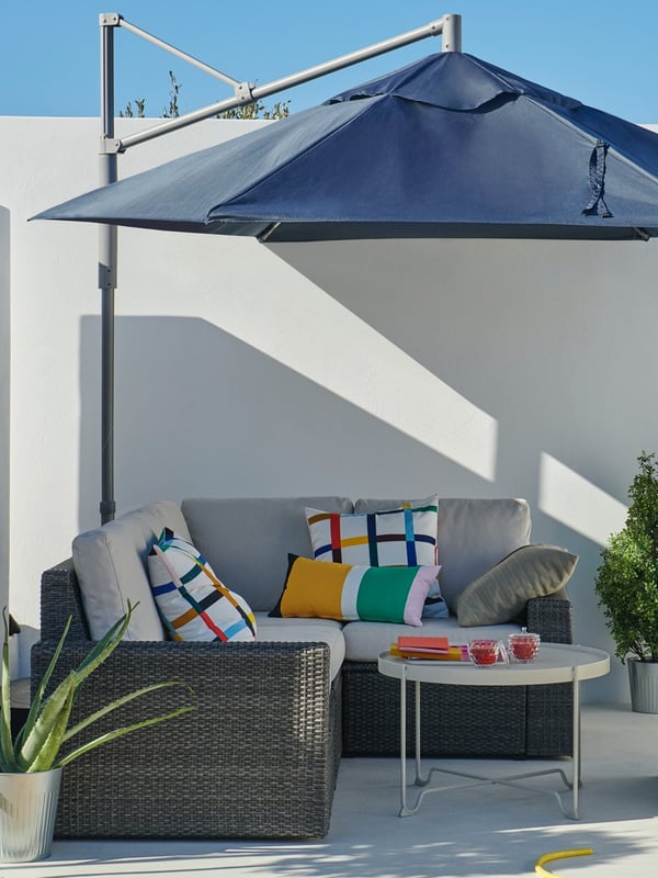 Un quitasol LINDÖJA cubre un living exterior con un sillon SOLLERÖN en L con cojines crema y rayados de colores, y una mesa de centro blanca KROKHOLMEN, frente a un muro blanco. 