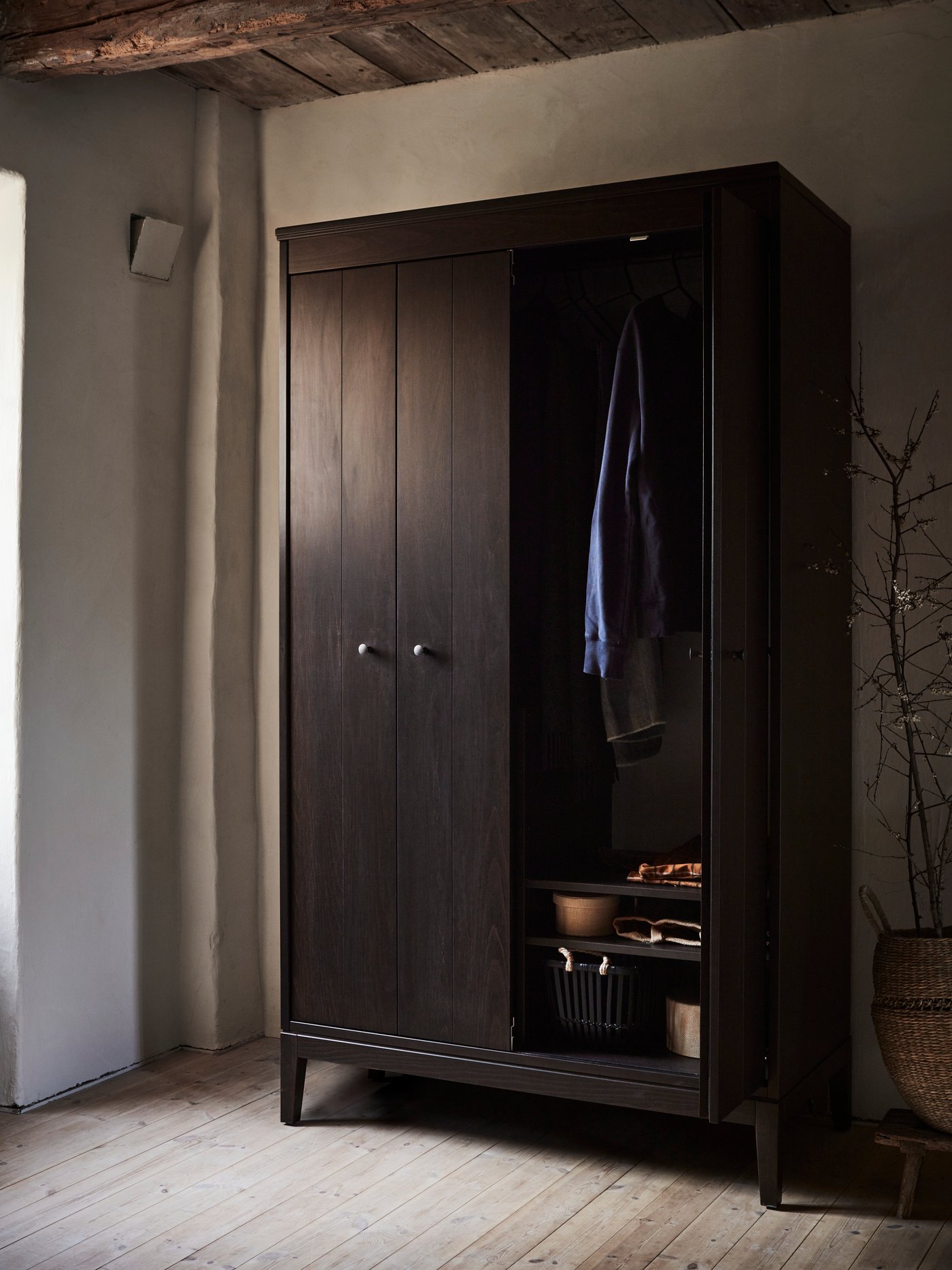 A dark brown stained IDANÄS wardrobe in a rustic style bedroom with the right hand door open and clothes on the hanging rail.