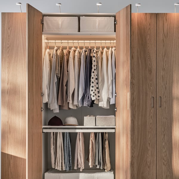 Un clóset PAX con puertas de madera con una puerta abierta para mostrar la ropa ordenada en su interior. Carrito negro con ruedas a la izquierda.