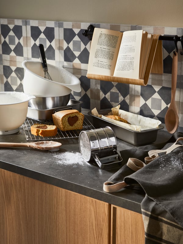 Una cocina con mesón EKBACKEN de color gris oscuro, con utensilios de cocina desordenados en medio de una preparación. El mueble base tiene puertas de madera. 
