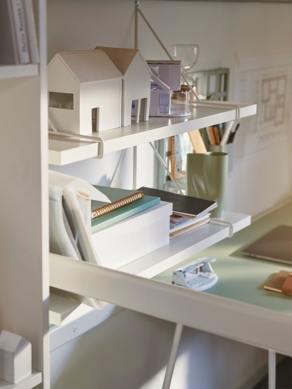 Two BERGSHULT/PERSHULT wall shelves with notebooks and little decorative houses painted white, hang on a wall above a desk.