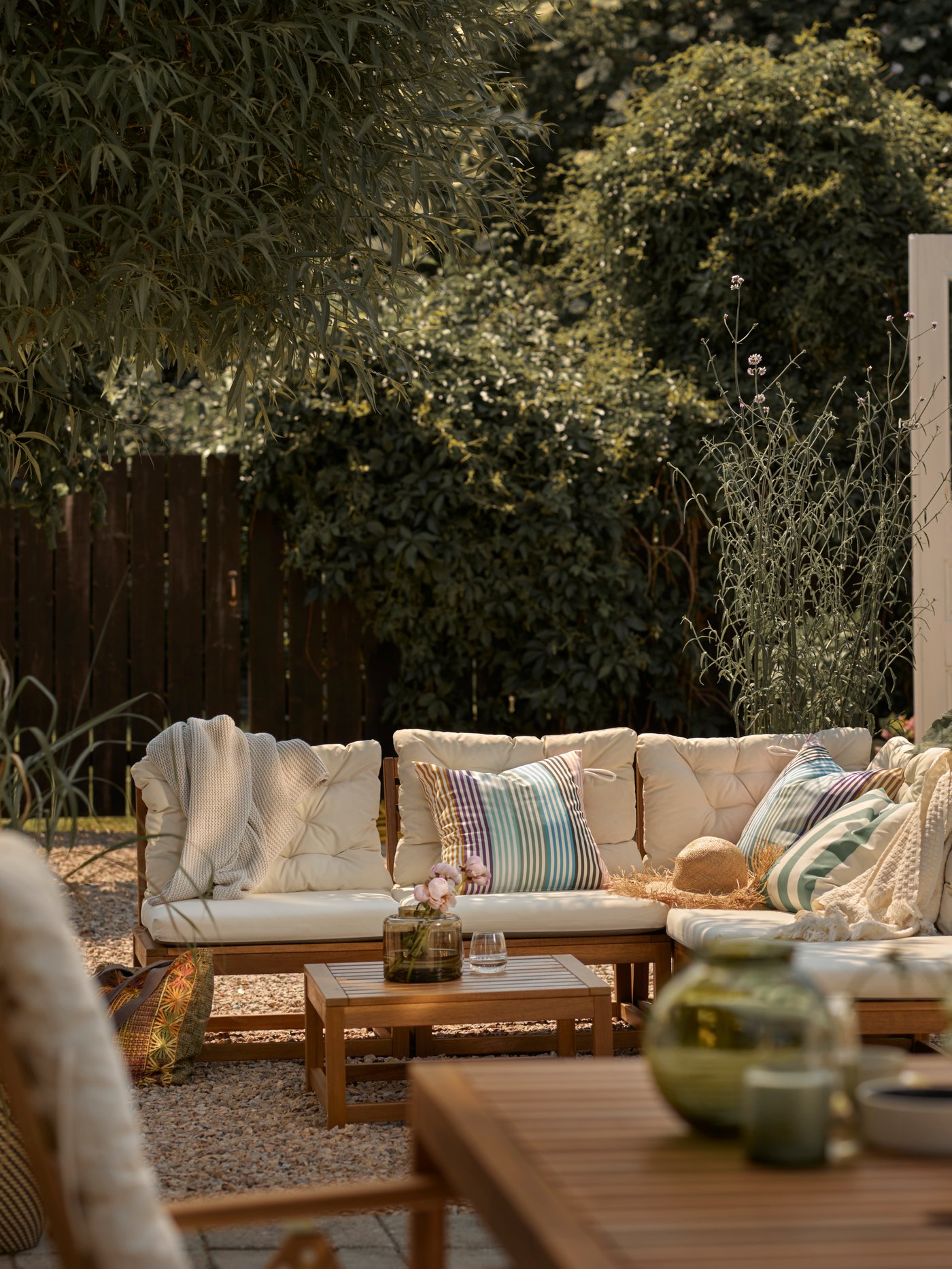 Un patio con árboles altos, en la terraza hay un set de living con muebles de madera que tienen cojines blancos.