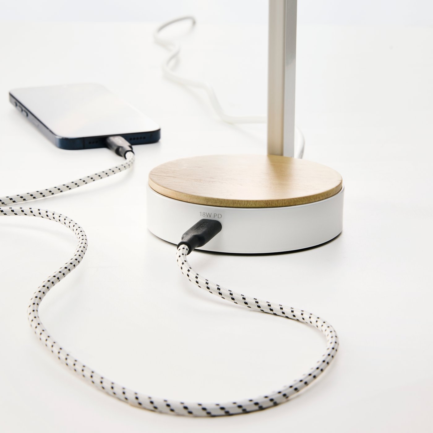 Et lyst skrivebord, hvor en mobiltelefon oplades, forbundet via et mønstret USB-C-kabel til en hvid ENSMÄRKE skrivebordslampe.