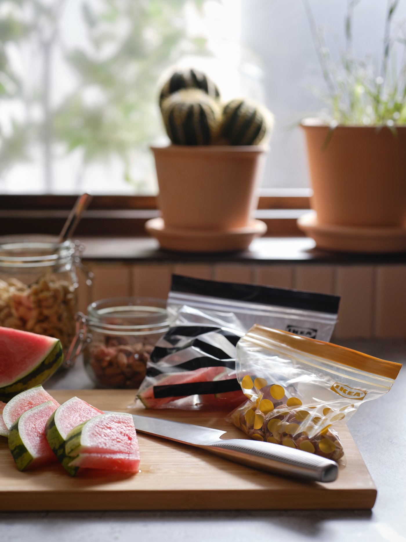 Slices of watermelon, a cook’s knife and two ISTAD resealable bags on a chopping board beside two clear glass jars with lids.