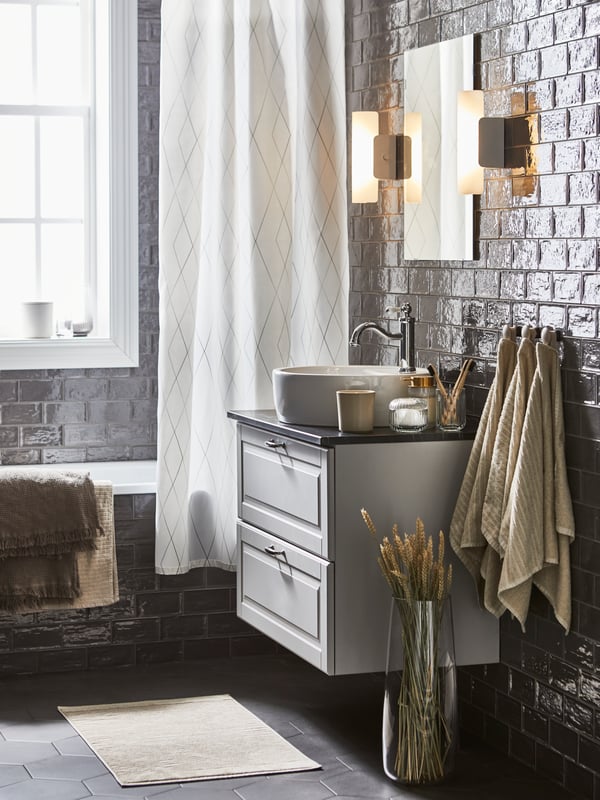 Une salle de bain avec un rideau de douche BASTSJÖN blanc/gris/beige, plus des serviettes et draps de bain brun/gris clair et beige clair.