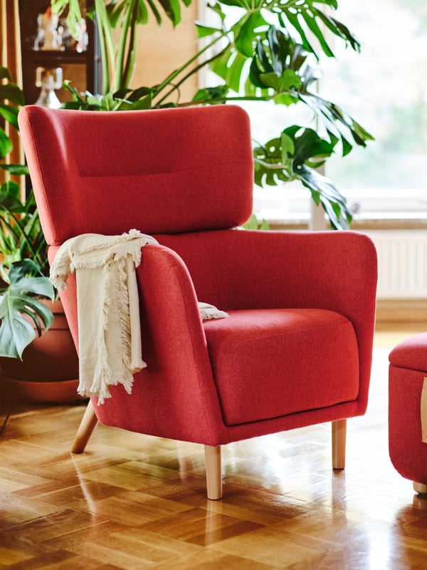 A red OSKARSHAMN wing chair with a red footstool and a beige blanket. A large lush green plant behind it in a living room.