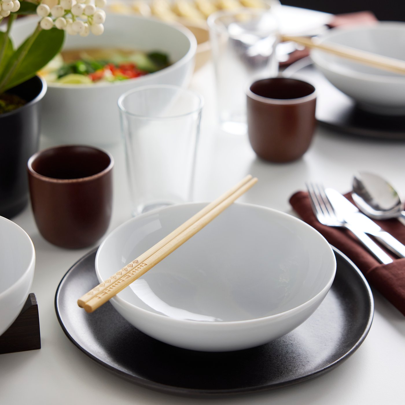 Two chopsticks lie on top of an IKEA 365+ bowl which stands on a black STOCKHOLM 2025 plate near an IKEA 365+ glass.