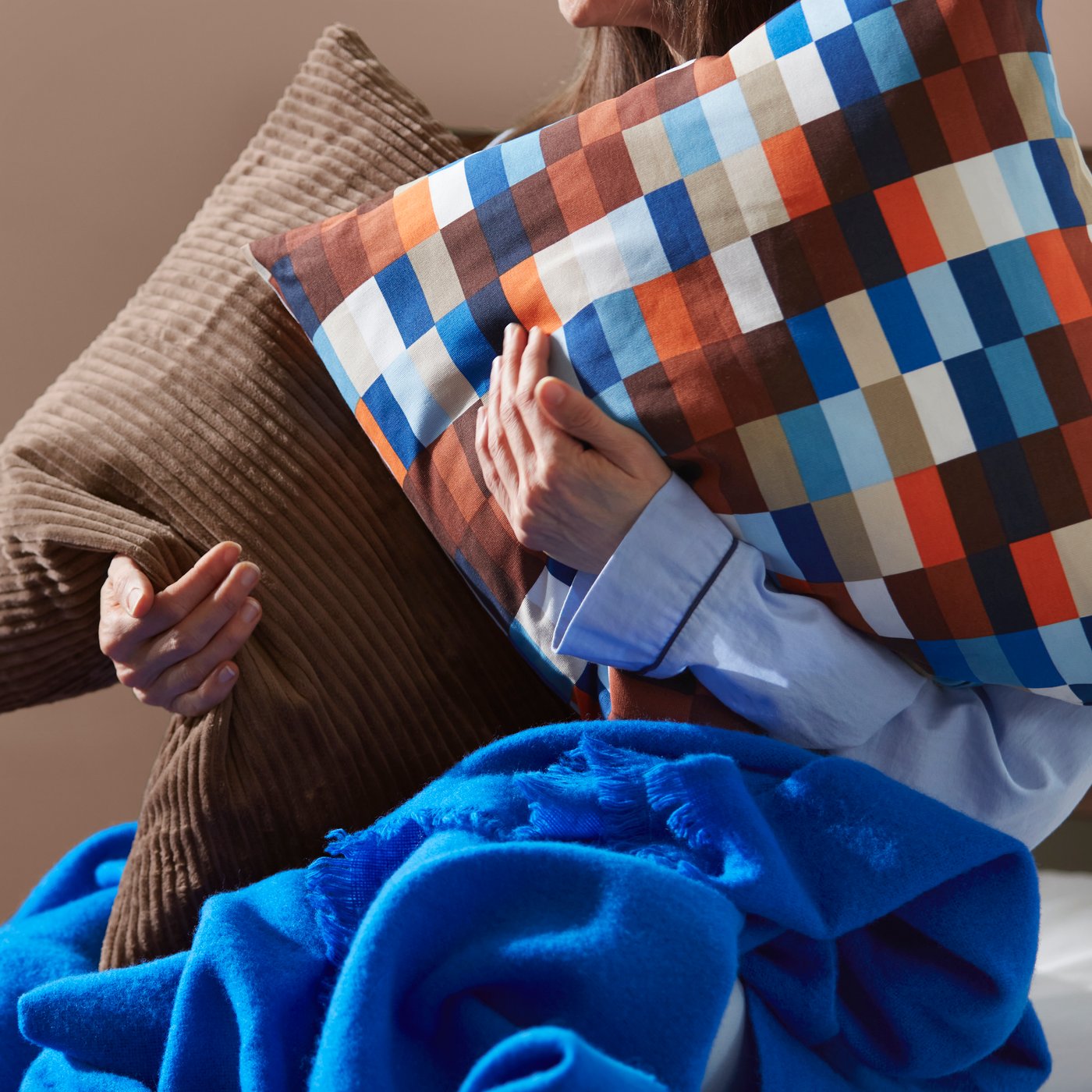 Eine Person hält zwei Kissen in der Hand; eines davon hat einen braun-blauen LÖVNATURSKOG-Kissenbezug mit bunten Quadraten.