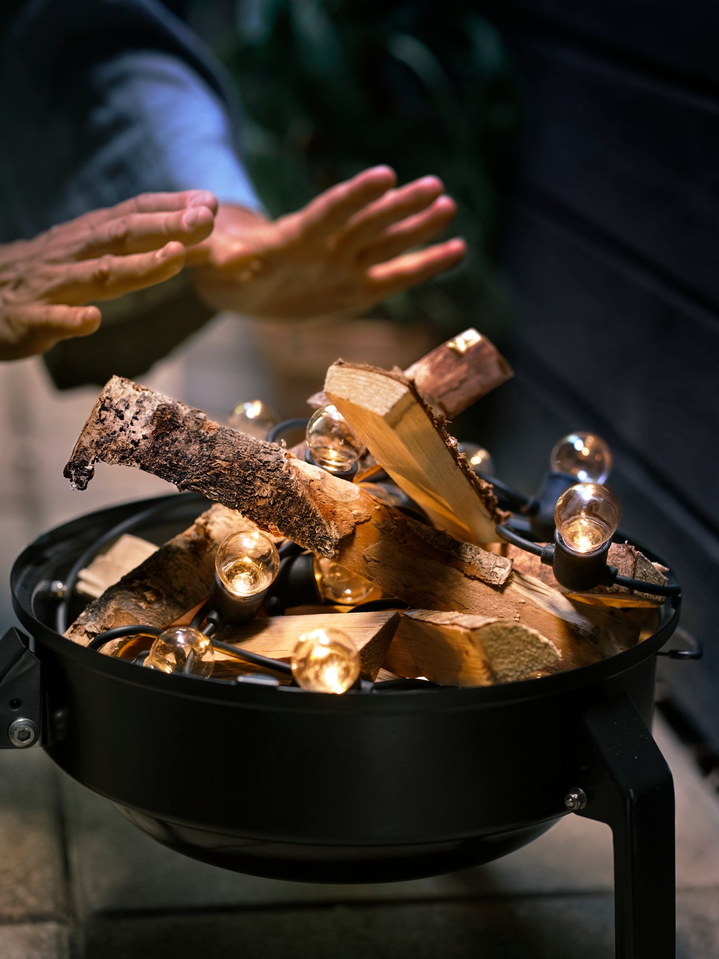 A pair of hands warming over a fake bonfire made of firewood and a black UTSUND lighting chain inside a portable barbecue.