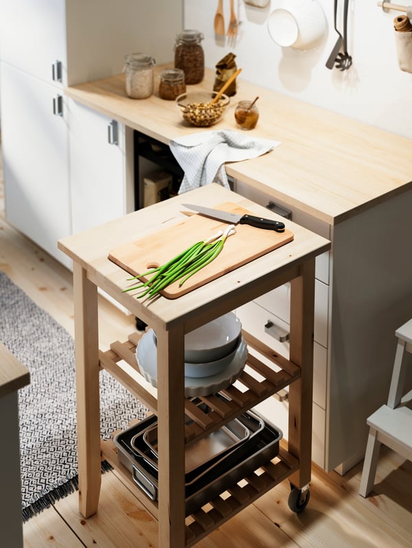 Green onions and a knife on a chopping board on a BEKVÄM birch kitchen trolley placed perpendicular to an EKBACKEN worktop.