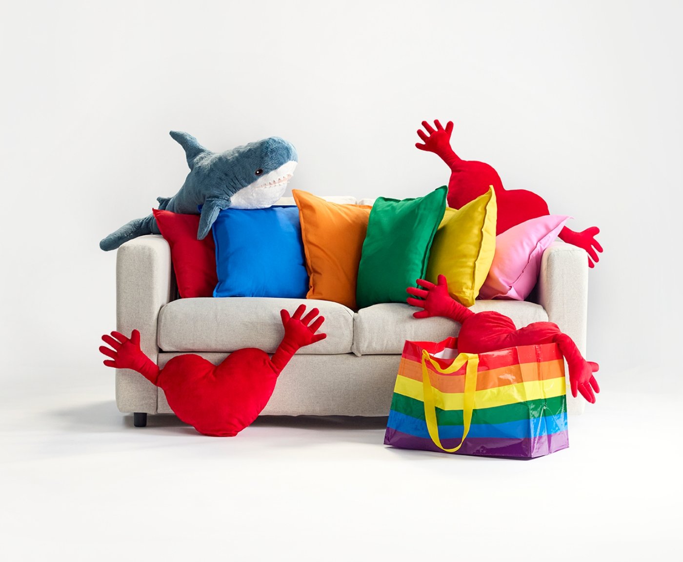 A beige sofa with several items on it: six GURLI cushions in red, blue, orange, green, yellow, and pink, two FAMNIG HJÄRTA cushions, and a BLÅHAJ shark soft toy. Next to the sofa is a rainbow STORSTOMMA bag containing a FAMNIG HJÄRTA cushion.
