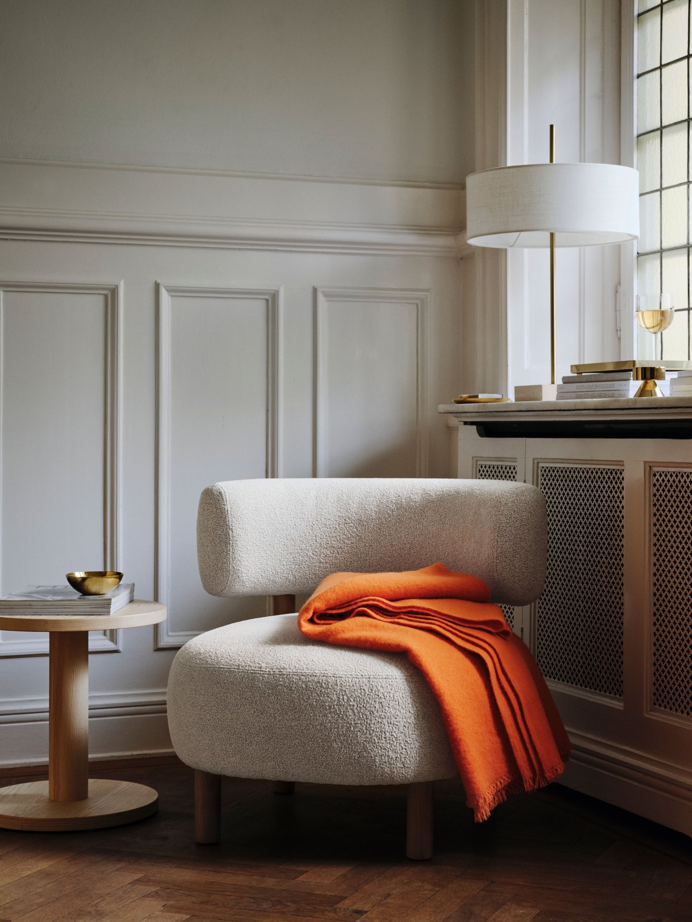 Una poltrona STOCKHOLM 2025 beige accanto alla finestra nell'angolo di una stanza. Un plaid arancione brillante è appoggiato sulla poltrona.