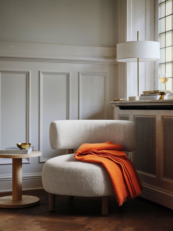 A beige STOCKHOLM 2025 armchair by a window in the corner of a room. A bright orange throw rests on the armchair.