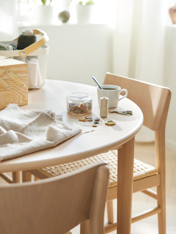 Dve SKANSNÄS stolice stoje za SKANSNÄS okruglim produživim stolom s RISATORP korpom, na kojoj se nalaze stvari za šivenje i šolja.