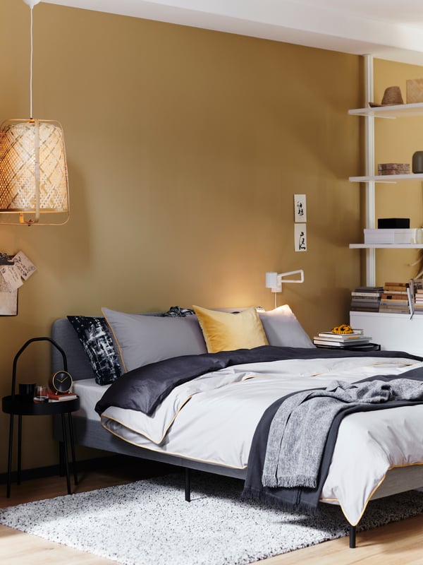 A grey HAUGA upholstered bed with a storage drawer pulled out, and a bedside table beside it holding a small table lamp.