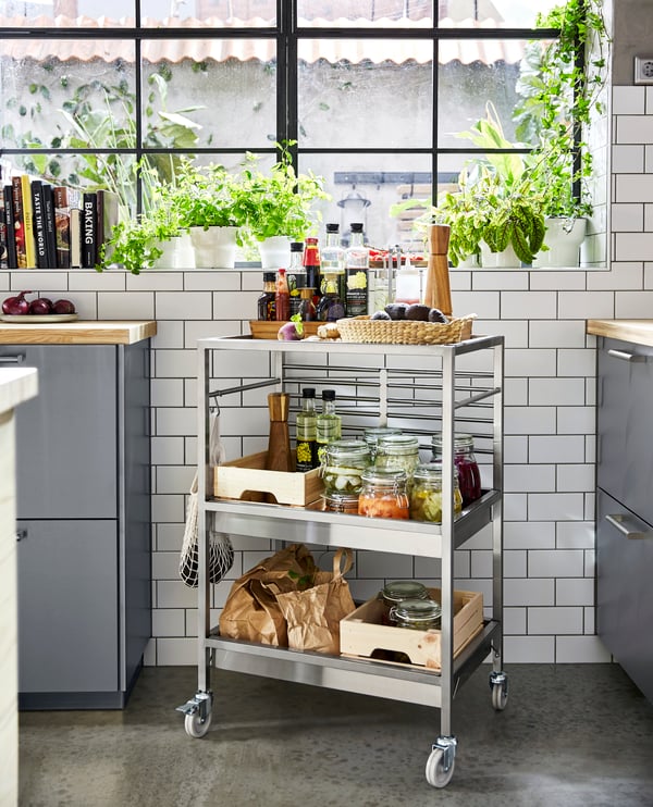 A stainless steel trolley on castors stands by a window. Oils, avocados and pickled vegetables are on the trolley shelves.