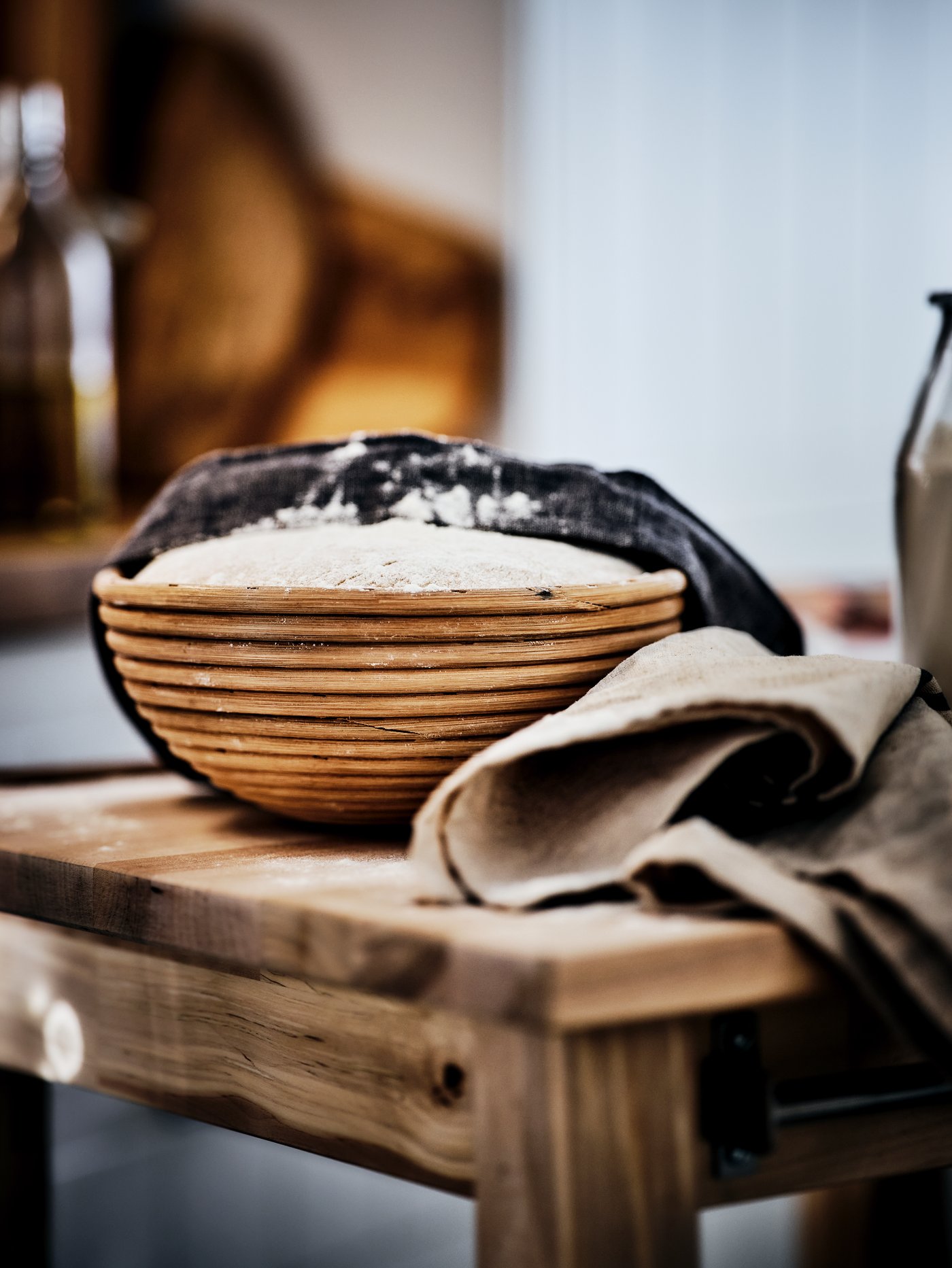 Une table en bois avec un banneton rempli de pâte à pain sur le dessus. Un linge à vaisselle recouvre à moitié la pâte.
