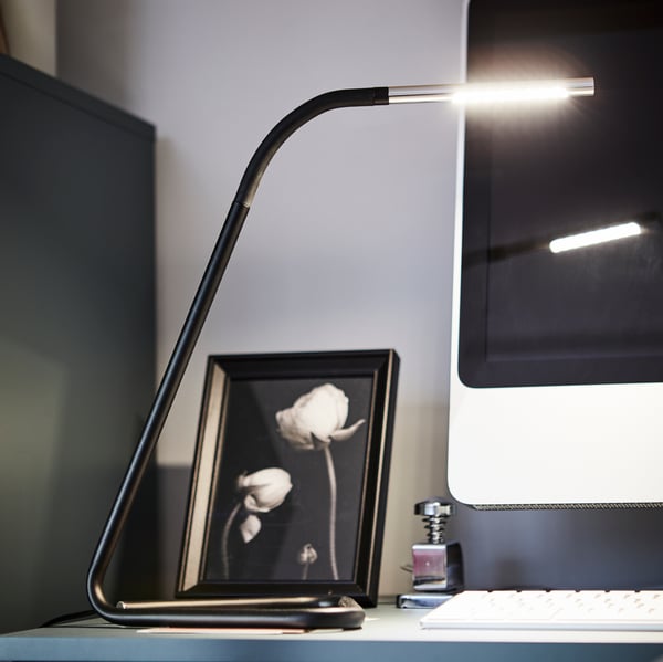 Lámpara de escritorio HÅRTE iluminando sobre un escritorio azul petroleo, en el fondo se ve un cuadro y una pantalla de computador.