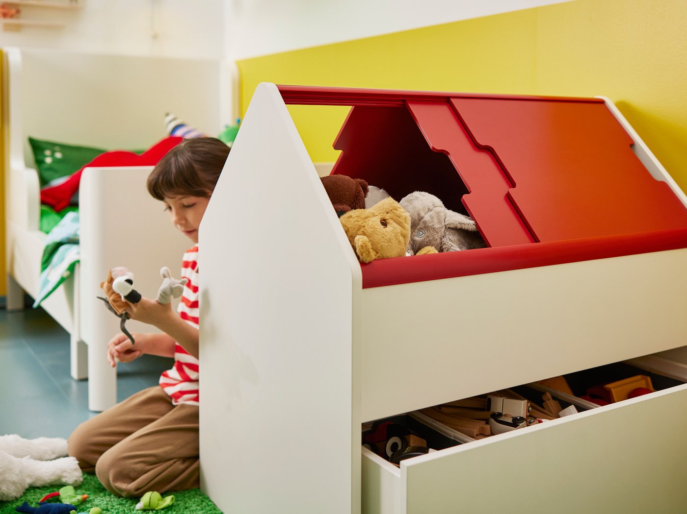 Une petite fille jouant avec des peluches, assise derrière un rangement VITBJÖRN rempli de jouets. Un lit évolutif BUSUNGE en arrière-plan.