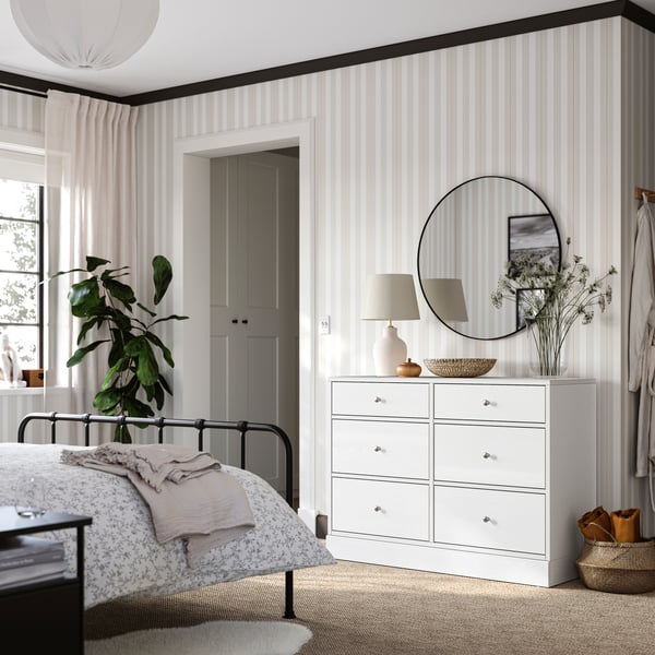 Chambre avec cadre de lit STJÄRNÖ, papier peint beige et blanc et commode HAVSTA blanche surmontée d’un miroir rond.