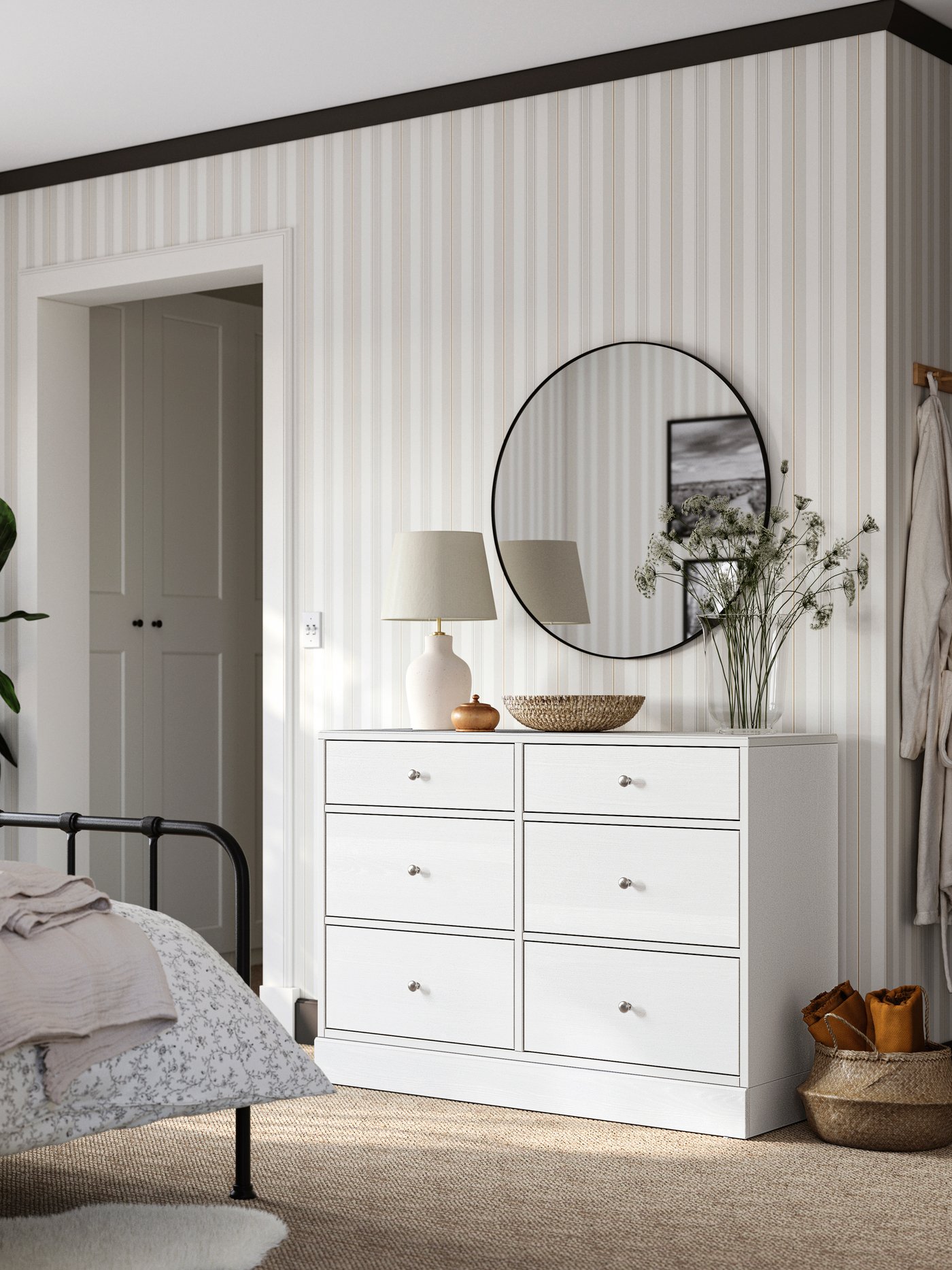Une chambre avec une structure de lit STJÄRNÖ, un papier peint beige et blanc et une commode HAVSTA blanche surmontée d’un miroir rond.