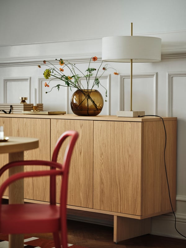 A brass-plated and white STOCKHOLM 2025 table lamp sits next to a brown vase on a cabinet by a white wall with wooden panels.