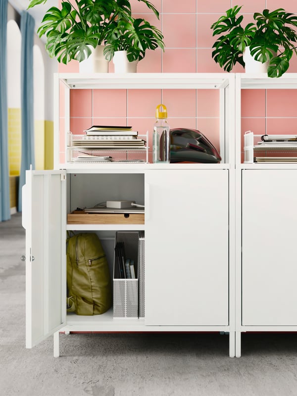 Side-by-side white TROTTEN cabinets with doors standing by a pink tile wall, holding leafy plants and office accessories.