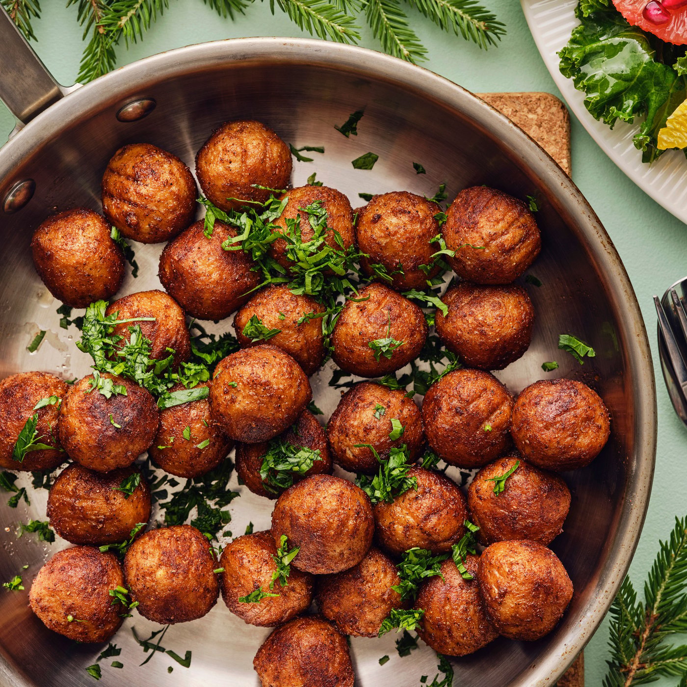 A frying pan filled with golden-brown HUVUDROLL plant balls garnished with fresh herbs is placed on a pale green surface.
