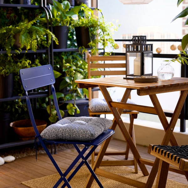 A blue KLÖSAN outdoor chair cushion with a floral pattern is tied onto a blue metal folding chair in a plant-filled balcony.