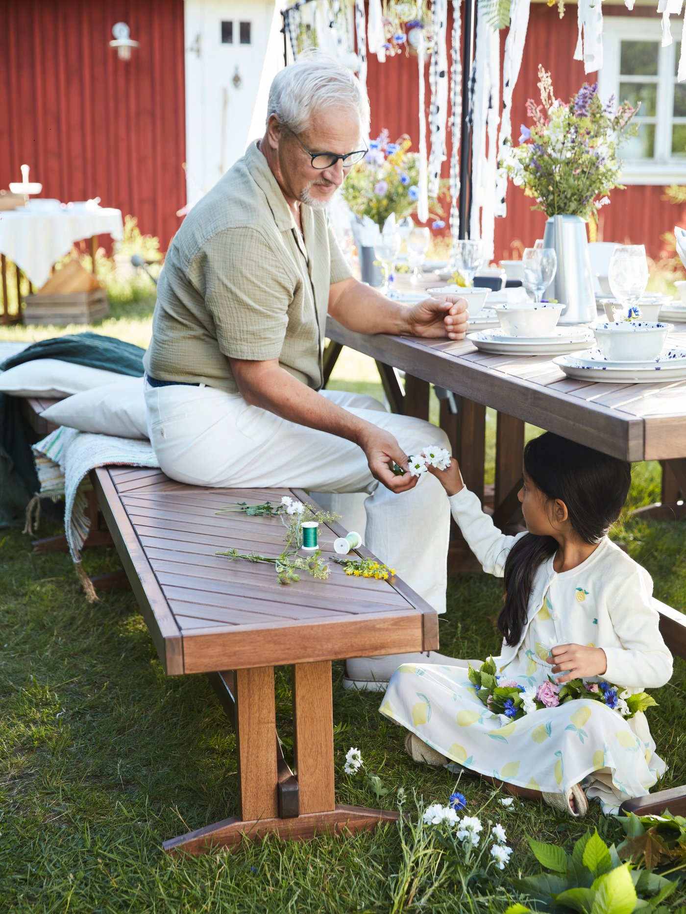 Ein Großvater sitzt an einem für ein Gartenfest dekorierten Tisch im Freien, während ein Kind darunter sitzt und einen Mittsommerkranz bindet.