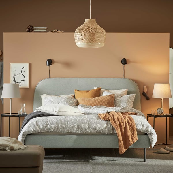 In front of a light brown bedroom wall is a curved, light green VADHEIM upholstered bed frame with many pillows.