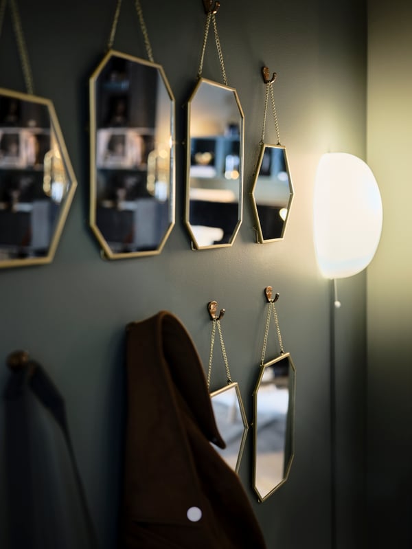 Rows of gold-colour LASSBYN mirrors hang on a green wall amid coats on hooks and a lit wall-mounted lamp.