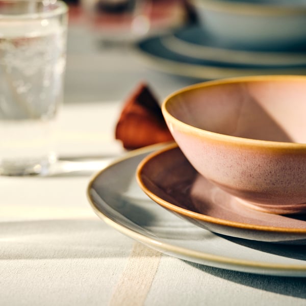 A table set with FÄRGKLAR bowls and plates stacked on top of each other, and a glass behind, all on a table mat.