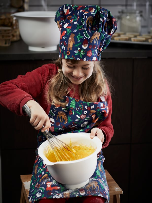 Una niña bate un cuenco con ingredientes húmedos para hacer masa de galletas. Lleva un delantal y un gorro de cocinero con motivos festivos.