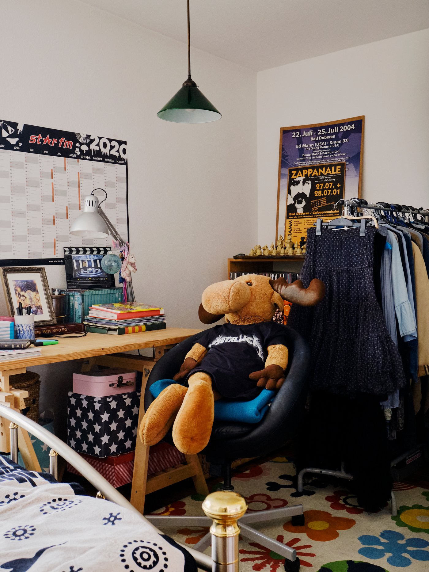 A large moose stuffed animal sits on a chair with clothes on in a room filled with decor and clothes.