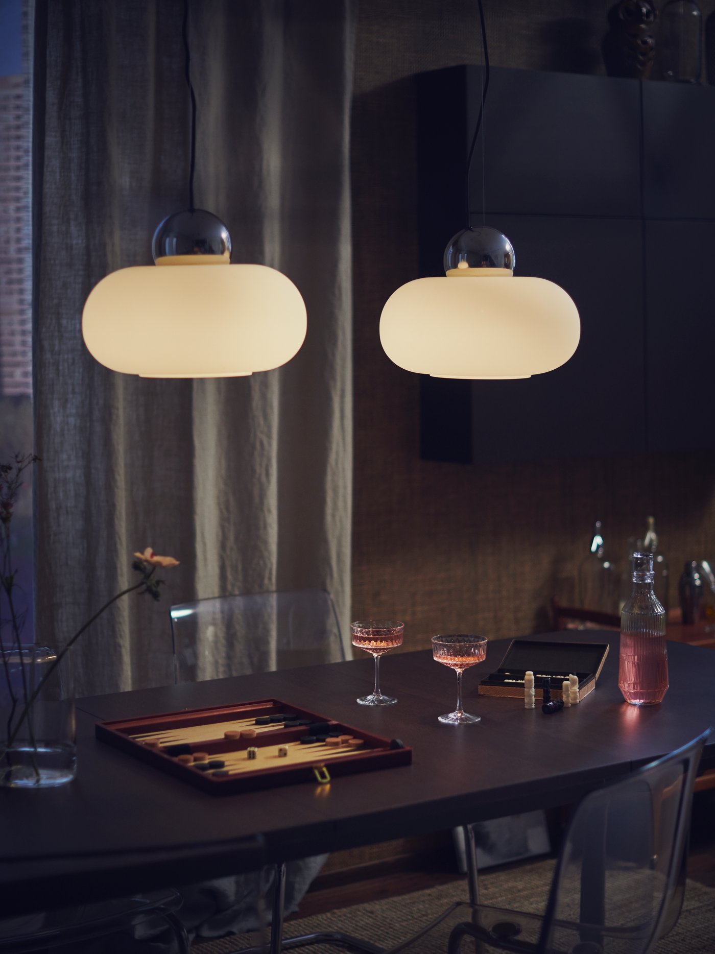 A cosy room at night with a board game and two glasses of wine on a table lit by two DEJSA pendant lamps.