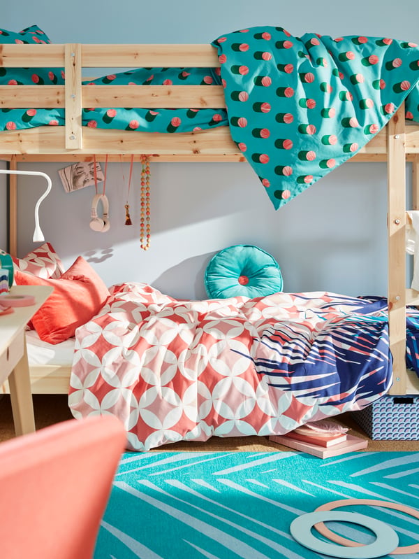 Un camarote MYDAL de madera natural apoyado en una pared celeste claro, las camas tienen ropa de cama de colores GRACIÖS y se ven desordenadas con objetos en uso. 