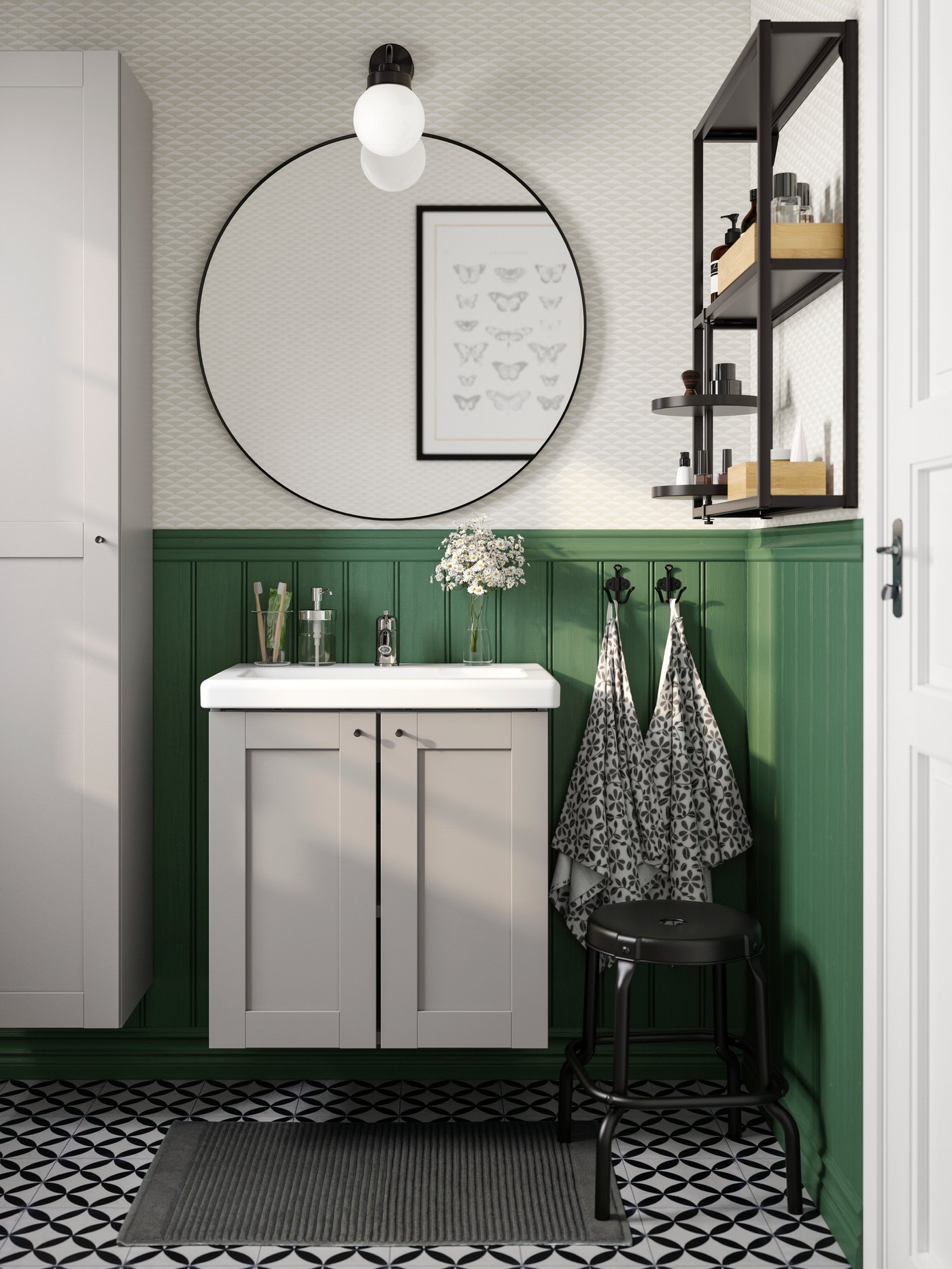 A big round black mirror hangs above a white wash-basin with two grey cabinet doors below it against a green wall panel.