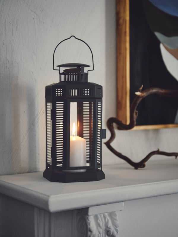 Lilin yang dinyalakan di dalam lantera lilin blok ENRUM berwarna hijau tua terletak di sebelah dahan yang berliku-liku di atas permukaan pendiangan hiasan.