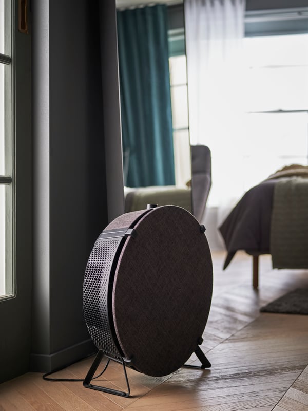 Close up on STARKVIND air purifier in black, standing on the floor with a mirror and a bed in the bedroom behind it.