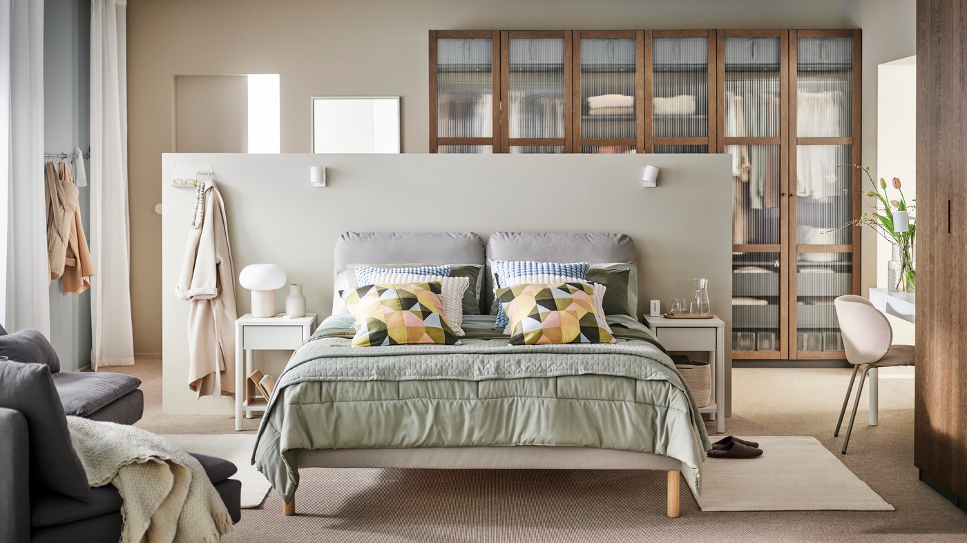 A brown SAGESUND upholstered bed in a beige bedroom with a grey-green KÖLAX bedspread and cushions in SVARTHÖ covers on top.