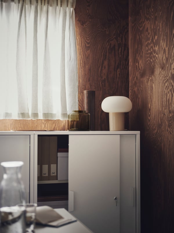 A brown wood-panel space. A white TROTTEN cabinet with sliding doors holds magazine files and has a DEJSA table lamp on top.