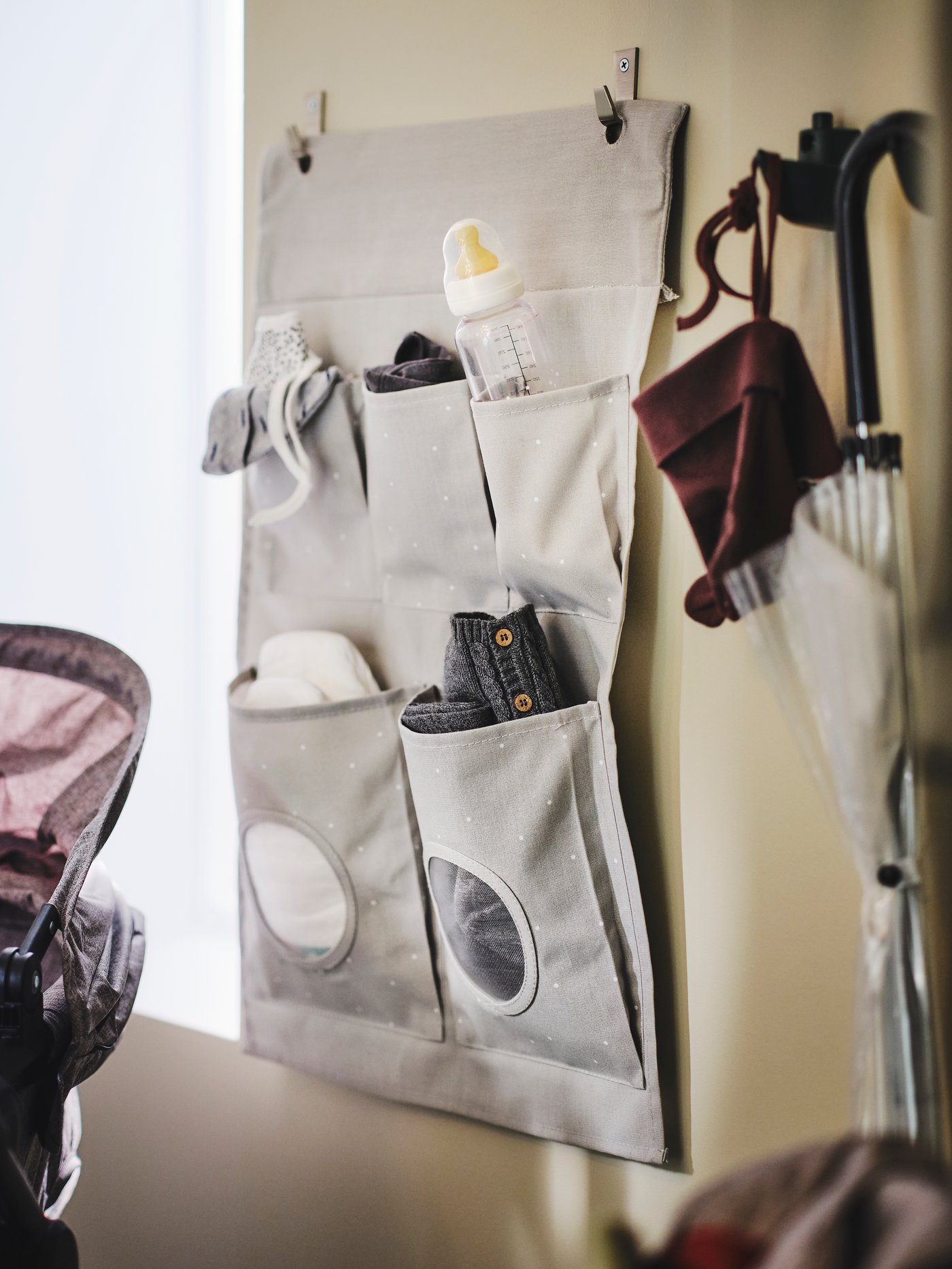 A dotted/light grey LEN hanging storage on a wall holding baby things like a bottle and nappies. A hat hangs on a hook by it.