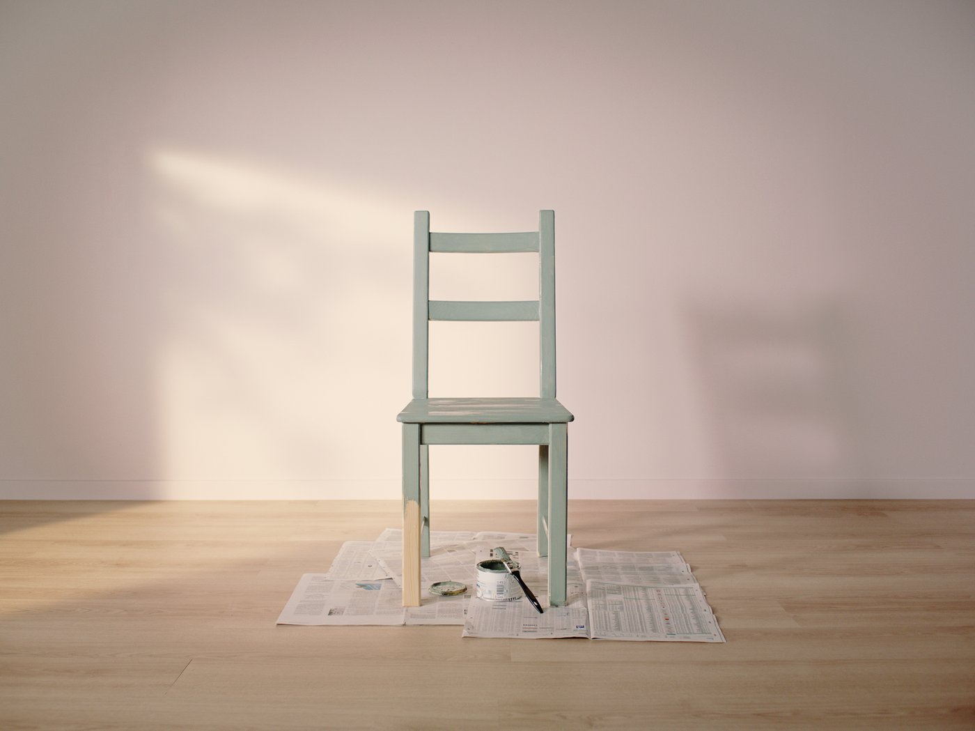 Une chaise IVAR peinte en vert clair posée sur de vieux journaux dans une pièce vide au parquet en bois clair et aux murs rose clair.