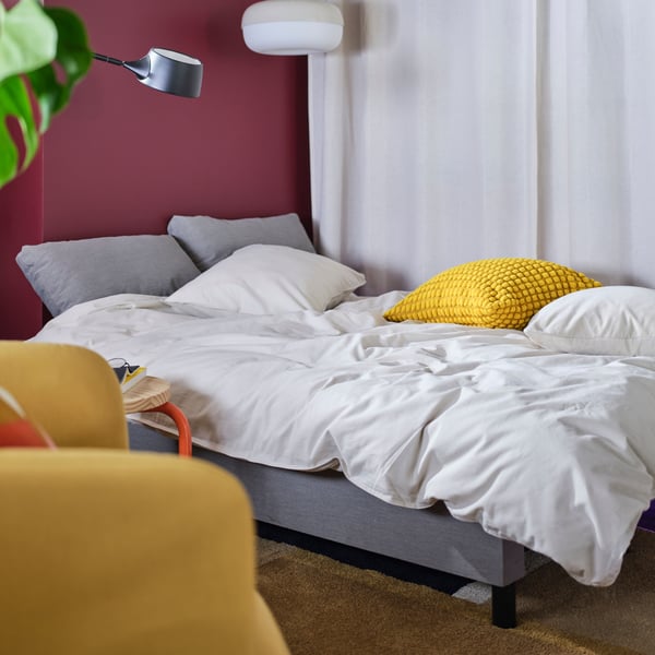 ÄLVDALEN sofa-bed in grey transformed as a guest bed with white bedlinen, a yellow pillow, a white pendant lamp and curtains.