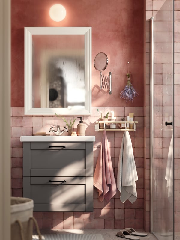 A bathroom with pink tiles has a grey ENHET washstand and a white mirror next to a shelf with hooks with two hanging towels.