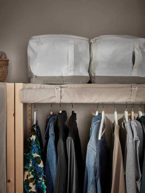 An minimal open wardrobe with fabric shutters. Inside a long line of clothes. On top two minimal fabric storage boxes. 