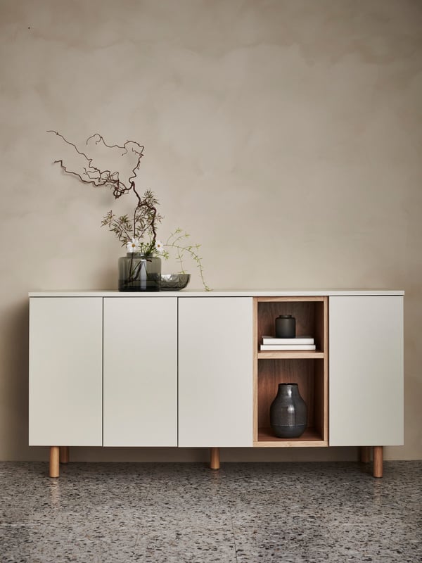 A LEDAMOT cabinet in grey-beige against a wall with a glass vase with flowers on top and a ceramic vase in the open section.