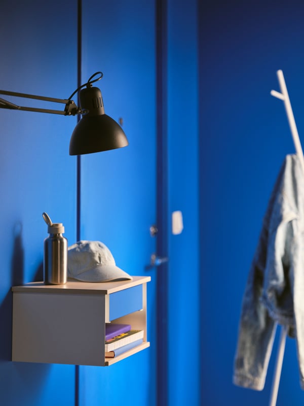 A STOMSÖ wall-mounted bedside table attached to a blue wall with a cap and a water bottle on top and a work lamp above it.