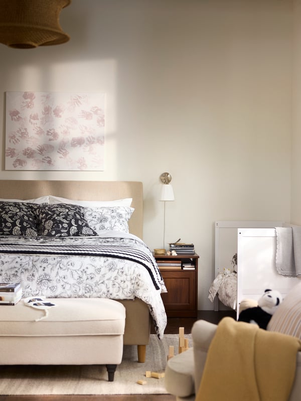 Schlafzimmer in Beige und Weiss mit einem Kinderbett neben dem Bett der Erwachsenen und einem Sessel im Vordergrund
